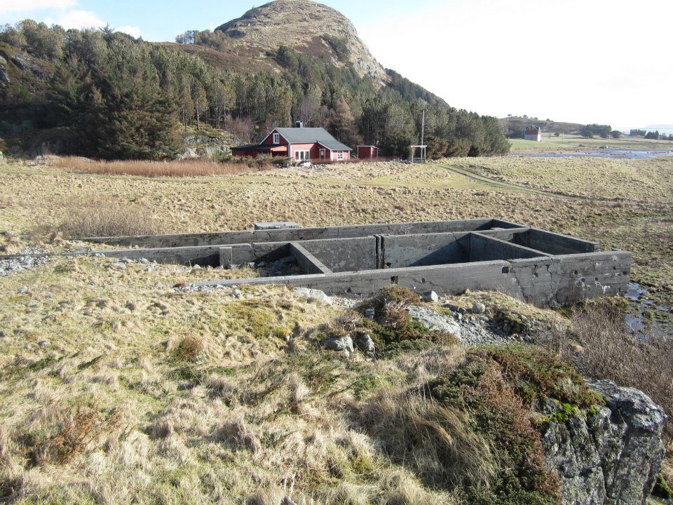 Grunnmurene etter anleggets største brakke. Rullestein-stranden like ved, som er fredet har &quot;slukt&quot; deler av grunnmuren. Huset i bakgrunnen var opprinnelig en brakke som var ibruk som lager og verksted. Like bak denne bygningen ligger der en bunker som er delvis innskutt i fjellveggen.